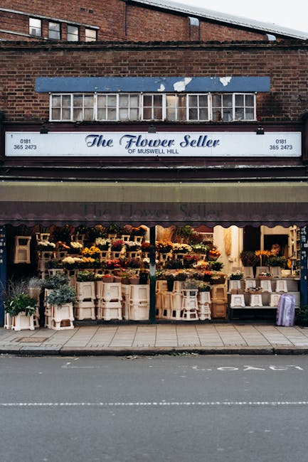 The flower shop, named 'The Flower Seller of Muswell Hill,' features a large storefront with a black awning and a white sign displaying the shop's name and contact number. The display window showcases