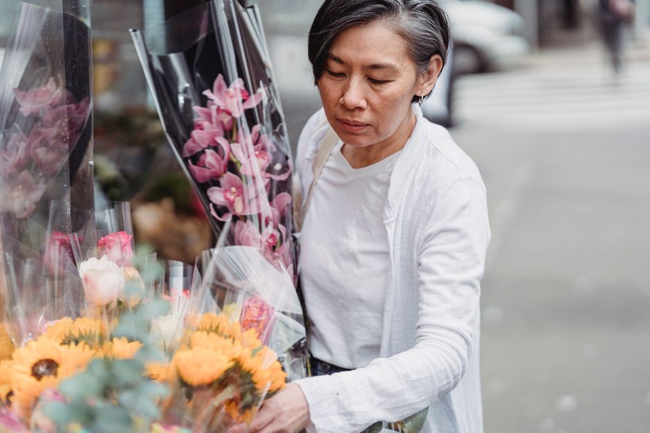 An older woman with short gray hair, wearing a white shirt and light-colored cardigan, examining a selection of fresh flowers at an outdoor flower shop, identifiable by vibrant bouquets of pink orchid