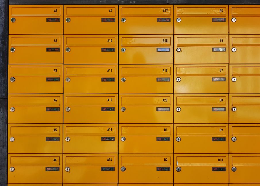 A collection of bright yellow postal lockers featuring multiple compartments with small, rectangular digital locks and keyholes, arranged in a grid pattern on a dark background. The lockers are unifor