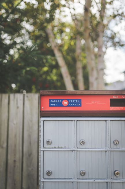 A close-up of a red Canada Post mailbox positioned outdoors in front of a wooden fence, with tall trees in the background. The mailbox features the Canada Post logo and various lock mechanisms, and is