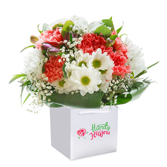 A masterful floral arrangement takes center stage within a pristine, square white gift bag, echoing the refined tranquility and vibrant greenery of Teddington. Plush carnations in shades of coral, peach, and vivid red display their abundant, ruffled petals, cascading in a soft, textured embrace. They are complemented by cheerful white daisy-like blossoms, each with a green-yellow center that recalls the delicate blooms found in the meadows beside the Thames. Airy sprays of white gypsophila are carefully scattered between the bold blossoms, imparting a gentle, cloud-like delicacy, while hints of purple-tinged buds emerge from behind, promising richness and layers. Lush, broad leaves in deep glossy green form a supportive base, wrapping the entire arrangement in a collar that mirrors the leafy parks stretching along Teddington Lock. Finished in a chic white bag with crisp ribbon handles for effortless carrying, the bouquet's rounded form suggests not only freshness and abundance but also the joy of local celebrations-be it a birthday, family milestone, or special thank you. The arrangement, expertly photographed against a seamless white background, brings the colours and intricate textures into sharp relief, creating a timeless image of local floristry at its finest, thoughtfully crafted to brighten the day of anyone receiving flower delivery in Teddington.