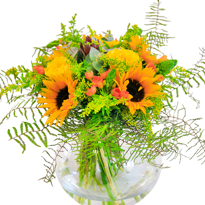 The first impression of this bouquet, designed near the heart of Teddington, is one of radiant, sunlit joy. Vivid yellow and orange sunflowers stand proud, their textured brown centers and beaming petals recalling the flower meadows scattered beside the Thames Path. Amidst these sunlight giants, yellow garden roses spiral open, their many-petaled blooms bringing a classic softness that balances the arrangement perfectly. Dotted among these are the glossy scarlet-red berries of Hypericum, shining with a playful energy that draws the eye. A cascade of ferns and flexing filler greens, soft and feathery, surrounds the main blooms-echoing both the gentle movement of the river breeze and the shading trees of local Teddington parks. Delicate yellow blossoms on slender spikes add further dimension, while subdued, blue-green eucalyptus leaves peek through, lending a subtle aroma and visual coolness. Displayed in a simple, round glass vase that glimmers with clear water, the stems intertwine in a pattern of natural beauty and careful curation. The arrangement brings to mind warmth, celebration, and openness-perfect for marking anniversaries, birthdays, or sending a cheerful token of affection, all thoughtfully crafted by local hands with a keen eye for Teddington's unique blend of natural charm and urban vibrance.