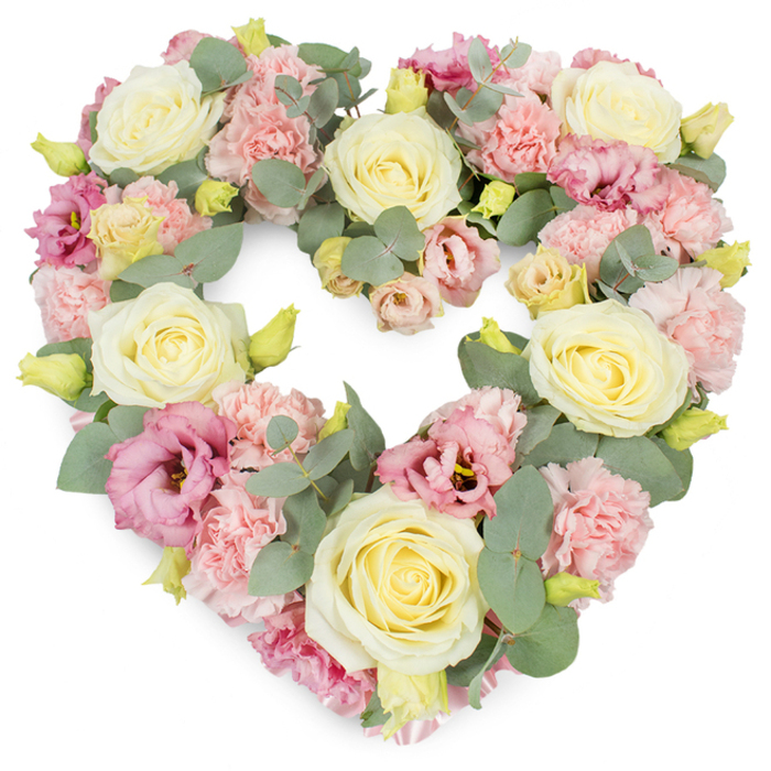 The photograph shows a large heart-shaped wreath, fully composed of pastel flowers and subtle greenery, placed against a spotless white backdrop. The heart is symmetrical and completely filled, creating a sense of richness and completeness. Pale yellow and creamy ivory roses are positioned strategically throughout the shape, each rose fully formed with tightly furled centres that spiral outward in velvety layers, suggesting both beauty and resilience. Nestled among these roses are numerous clusters of light pink carnations, their many ruffled petals overlapping to create soft mounds of texture that blur any harsh lines. Interspersed within the pastel mass are deeper shades of pink and mauve lisianthus, their more open petals and occasional darker centres forming gentle points of contrast and drawing the eye across the arrangement. Woven delicately through the flowers, silvery-green eucalyptus leaves provide a cool counterpoint, their rounded form and soft hue adding depth and a soothing, natural presence. Small yellow-green buds punctuate the design in places, giving quiet hints of growth and renewal. The whole heart feels lush and expertly balanced, reflecting careful craftsmanship. One can imagine this tribute being delivered to a farewell service near Teddington Lock or placed tenderly at a crematorium chapel frequented by local families. With its blend of roses, carnations, and lisianthus in creams, pinks, and greens, the wreath speaks of sympathy, enduring love, and cherished memories. It is a visually comforting piece, offering a serene focus for reflection during funerals, memorials, or anniversaries where remembrance takes centre stage.