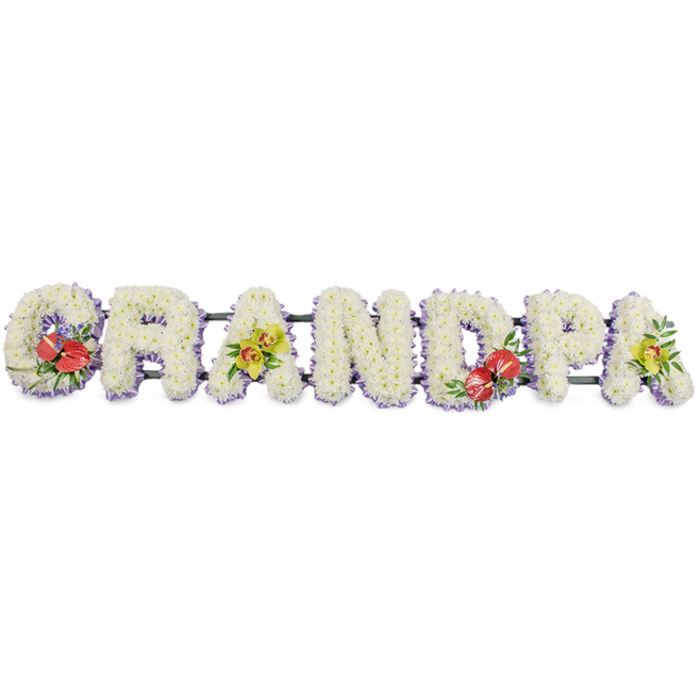 This photograph presents a personalised funeral tribute spelling "GRANDPA" in large, three-dimensional capital letters, arranged in a steady horizontal line against a bright white background that makes the design appear fresh and serene. Each letter is carefully packed with small white chrysanthemums, the petals overlapping to create a thick, textured surface reminiscent of soft quilting, offering a visual sense of comfort and calm. Along the lower edge of every letter sits a narrow band of pale lavender ribbon, peeking through just enough to form a delicate outline, adding definition and a touch of gentle colour without disturbing the tribute's predominantly white, peaceful palette. The letters are mounted on slim dark green bars that keep them aligned and secure, allowing the tribute to be displayed in one continuous piece at a chapel, crematorium, or graveside. Thoughtfully placed accent clusters provide contrast and character: on the "G" and the "D" are bold groupings of red anthuriums, their heart-shaped, waxy spathes and central spadices surrounded by glossy foliage and subtle filler, representing enduring love and heartfelt remembrance. The first and last "A" carry delicate yellow-green orchids, their graceful shapes and fresh leaves introducing an air of sophistication and hope. The tribute feels like a deeply personal message translated into flowers, the type of arrangement families across Teddington might commission for a grandfather who loved walks along the High Street or Sundays in Bushy Park. Every element, from bloom choice to colour placement, reflects the steady hand and empathy of an experienced local florist.